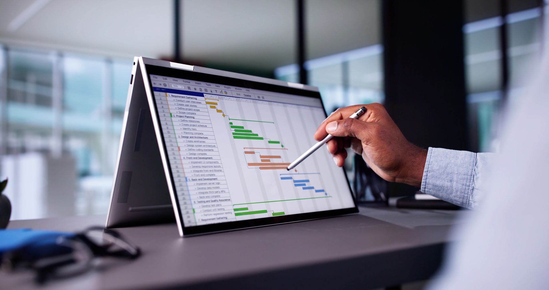 Person uses stylus to interact with a project timeline on a laptop in a modern office.