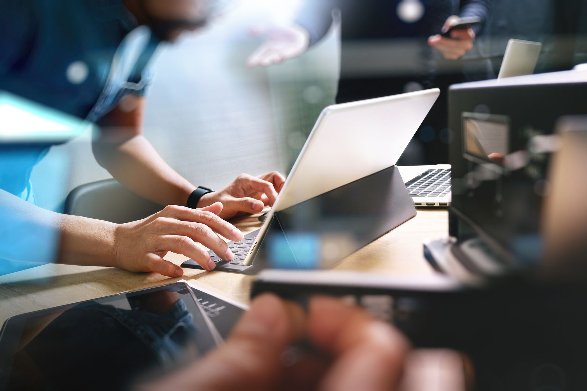 Hands typing on a laptop at a desk with other electronic devices and another person blurred in the background.