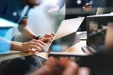 Hands typing on a laptop at a desk with other electronic devices and another person blurred in the background.