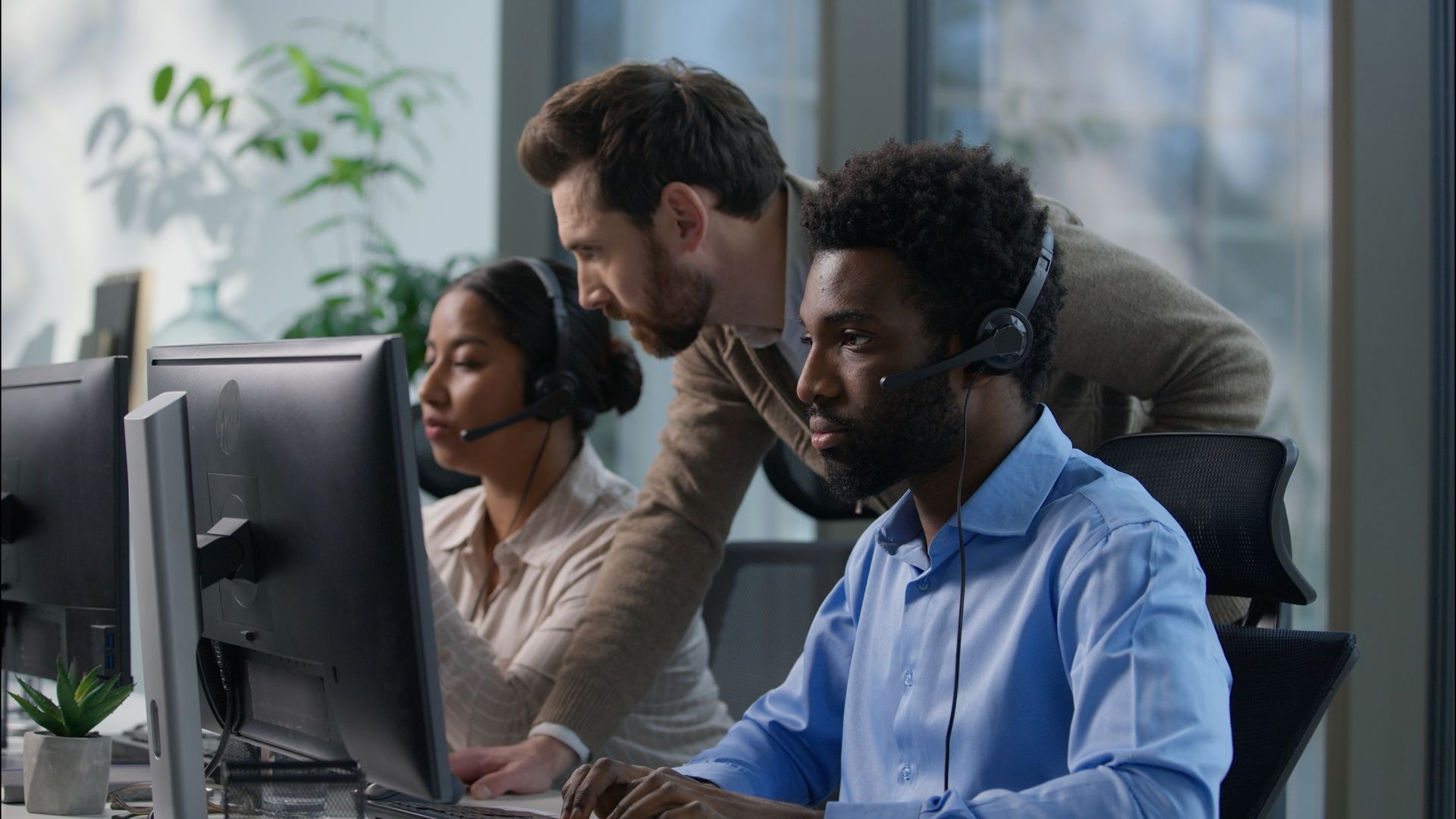 Three people in an office wearing headsets; one man leans over another's shoulder, pointing at a computer screen.