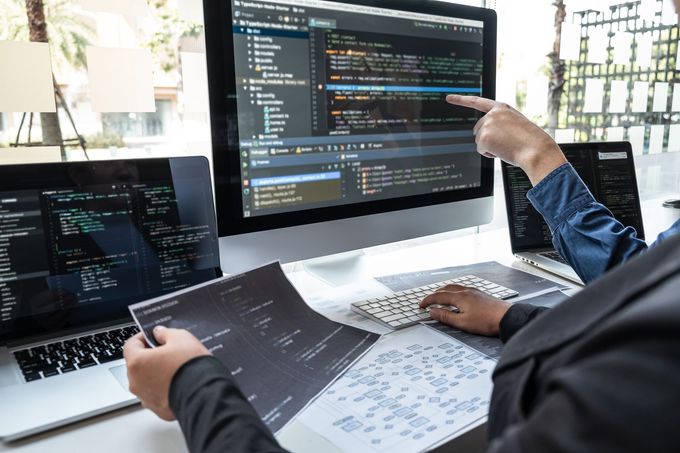 Person points at code on a computer screen, collaborating with others, possibly debugging.