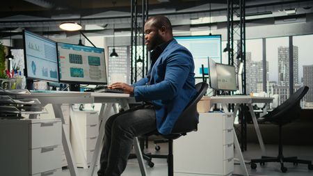 Man in blue jacket, working at computer in modern office with city view.