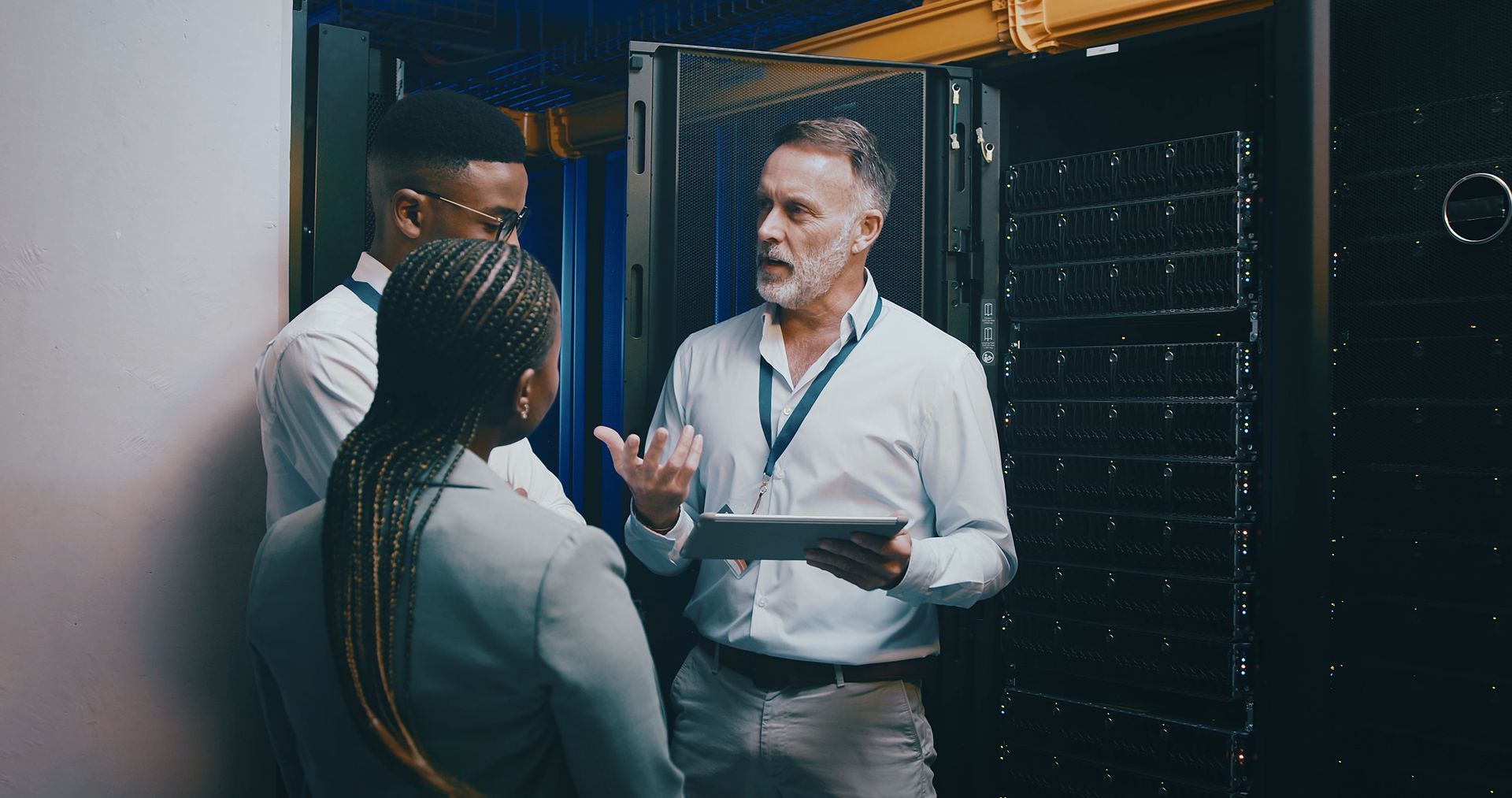 Three people in a server room, man holding tablet discussing with two colleagues near dark server racks.