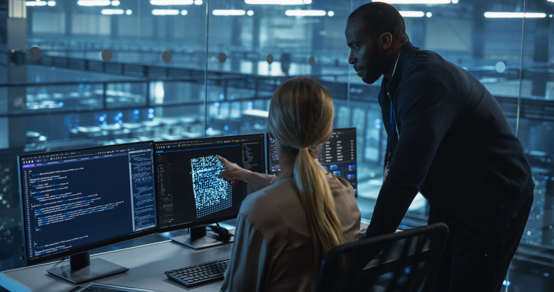 Two people at computer monitors, pointing at screen. Data center setting with blue lighting.
