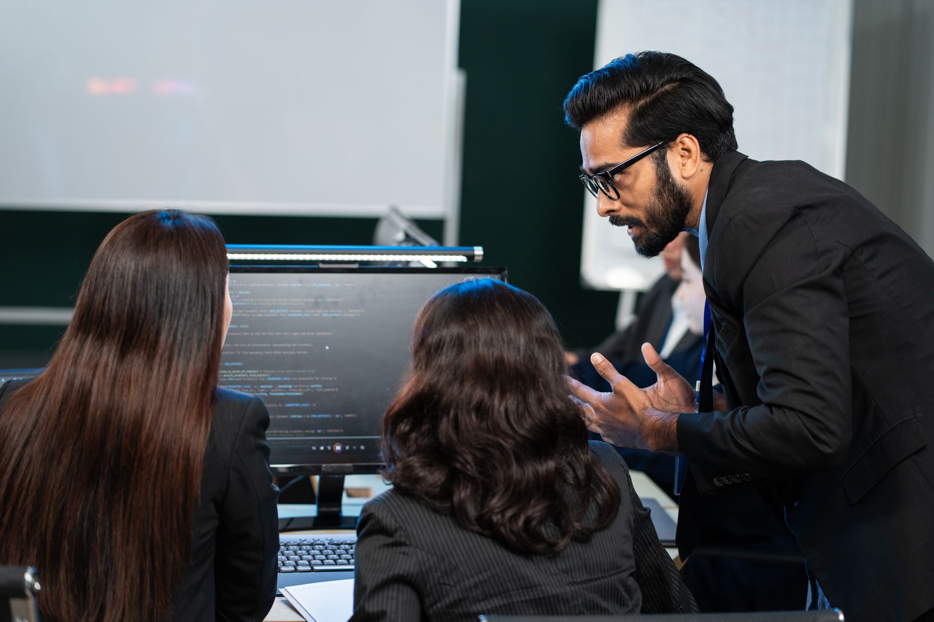 Man in suit gestures at a computer screen as two women look on. A workplace scene.