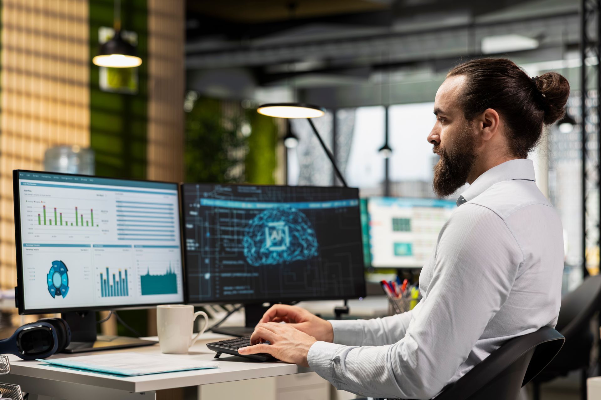 Man with a beard works on dual computer monitors in an office setting.
