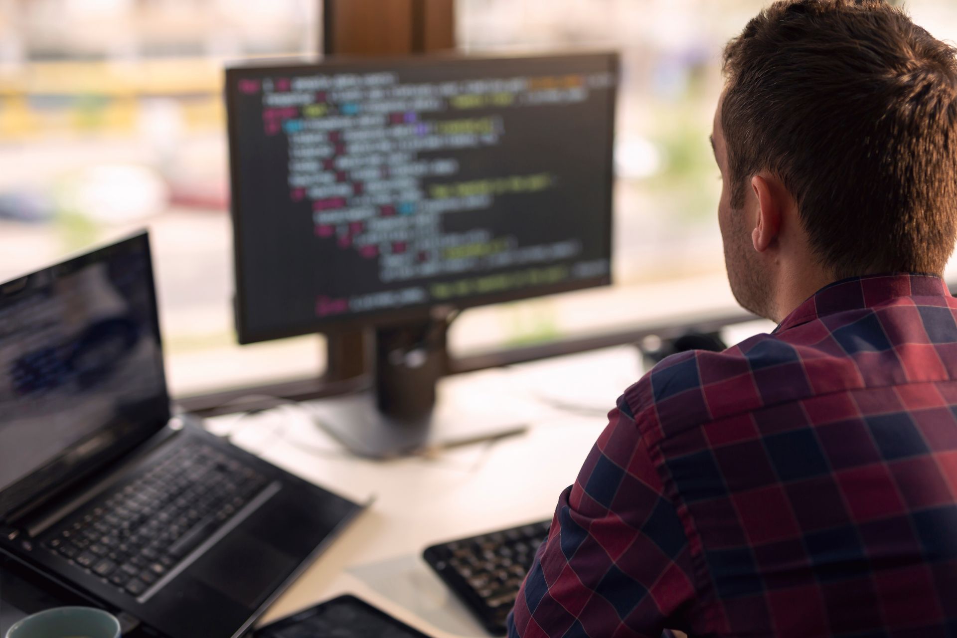 Person coding at a desk with a laptop, monitor displaying code, keyboard, and tablet.