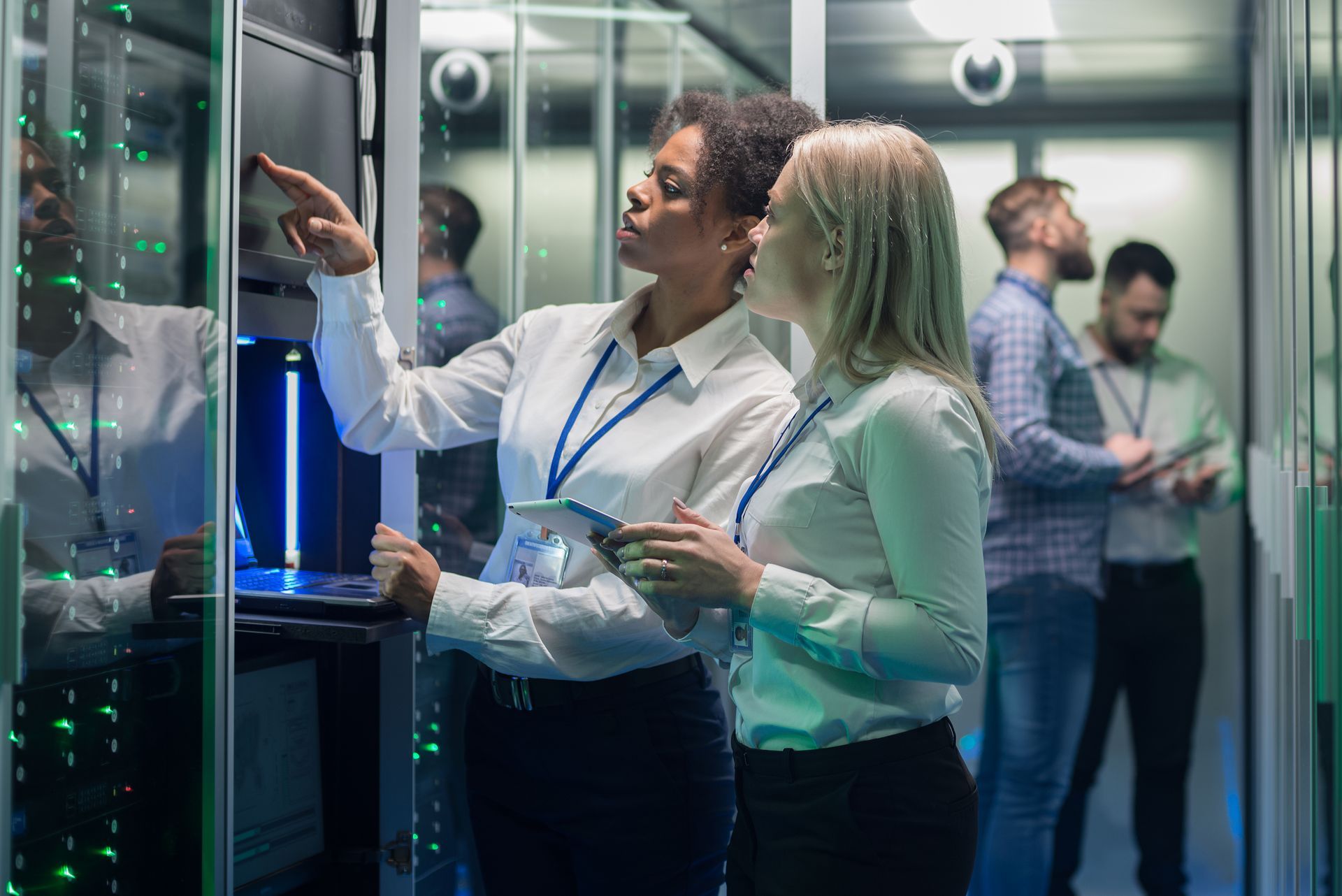 Two people in a server room looking at equipment. Other people are in the background.