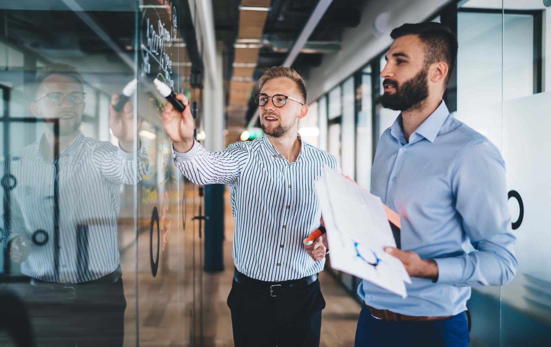 Two men in an office brainstorming on a glass wall; one writes with a marker, the other holds papers.