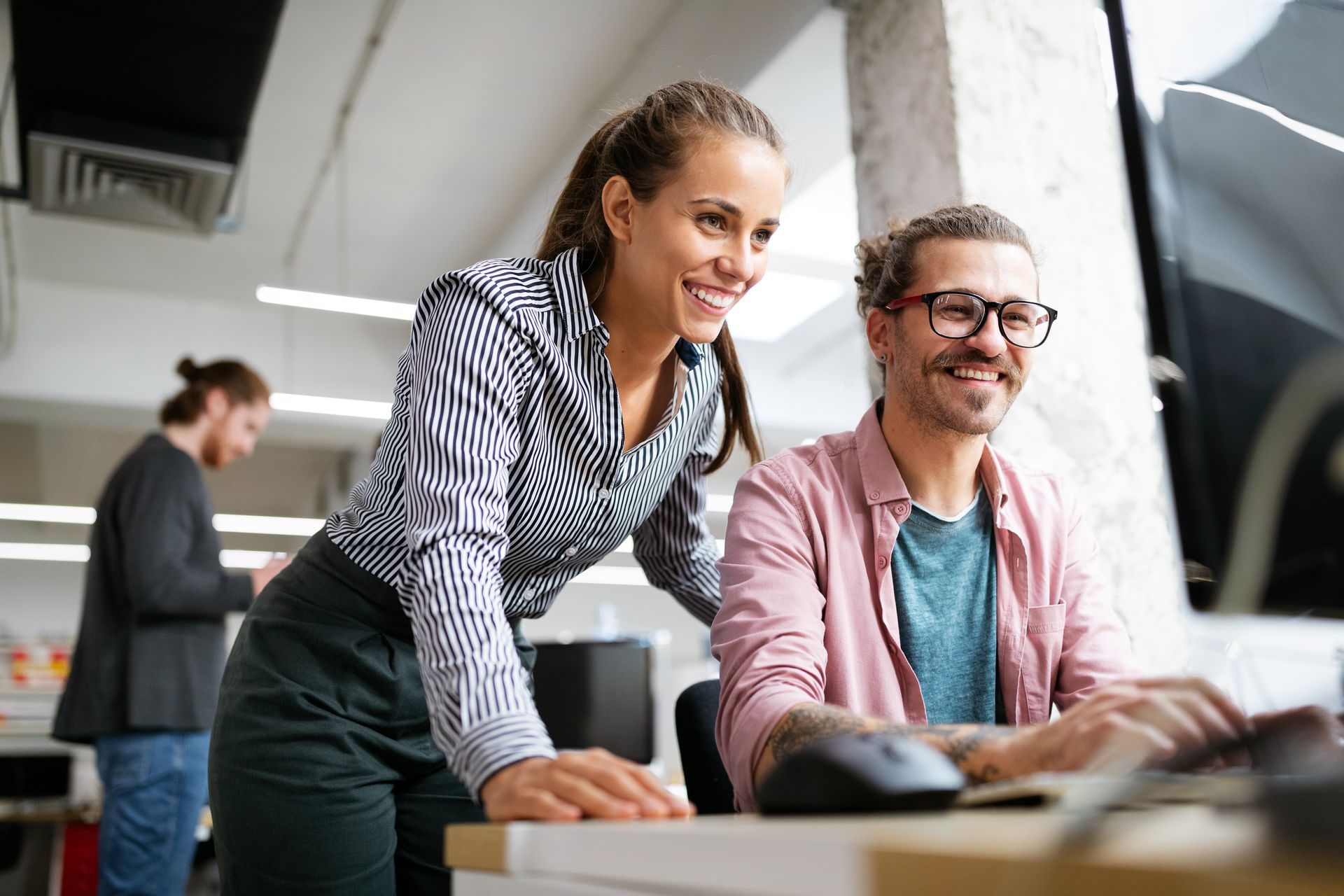 Woman and man smiling, looking at computer screen in office. Another person in background.