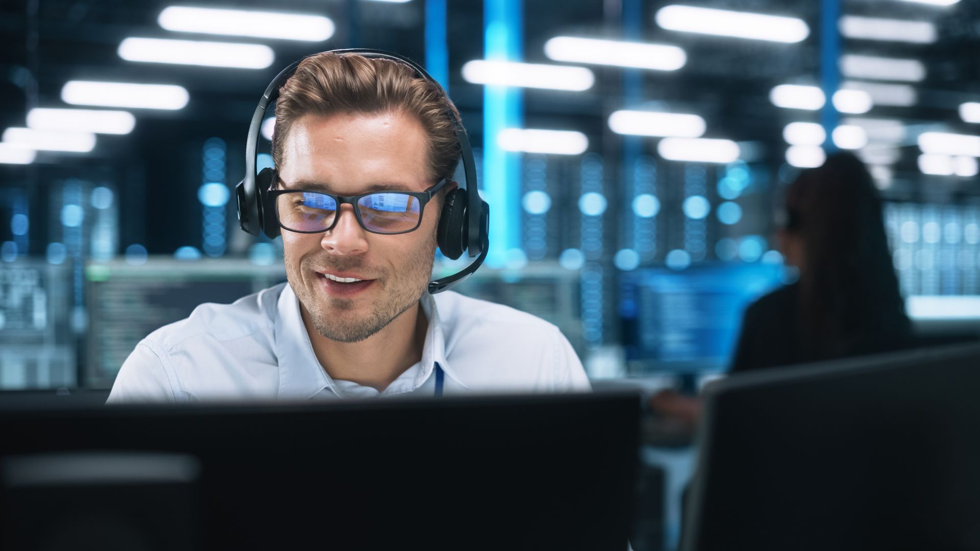 Man with headset and glasses smiling at a computer screen in a server room.