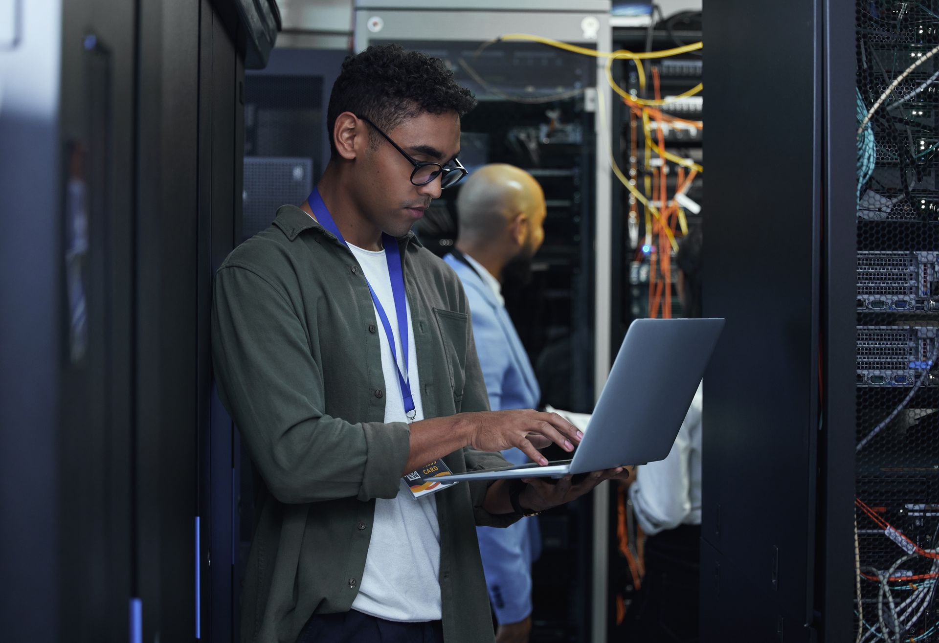 Man with glasses using a laptop in a server room, another person in the background.