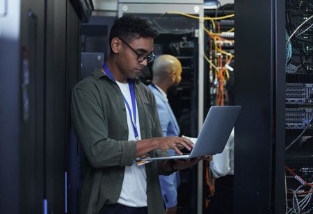 Man with glasses using a laptop in a server room, another person in the background.
