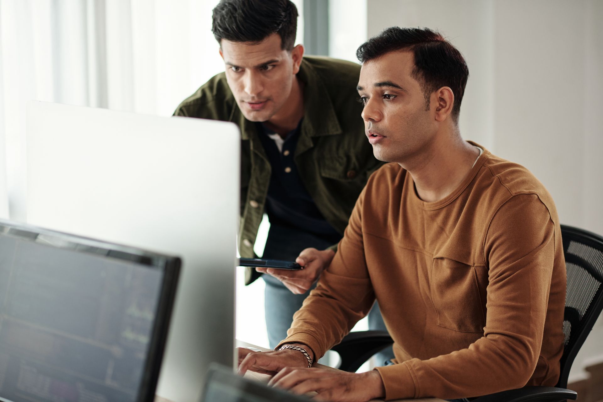 Two men looking at a computer screen in an office setting. One typing, the other looking on, both focused.