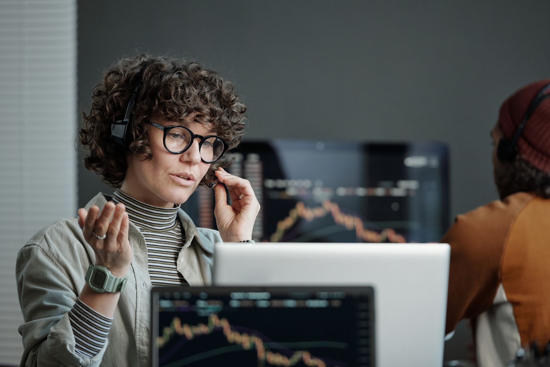 Woman wearing glasses and headset in front of a laptop, gesturing, with stock charts on the screen.