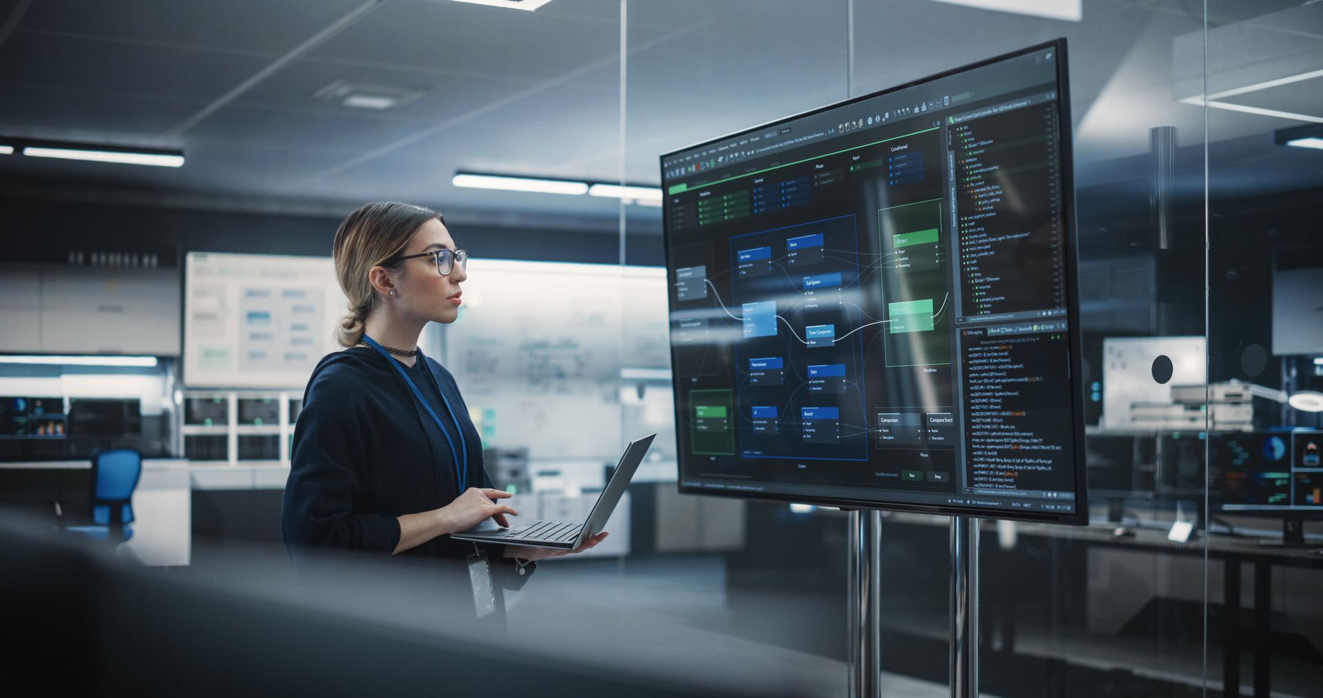 Woman with glasses and laptop examines data on a large screen in a modern control room.