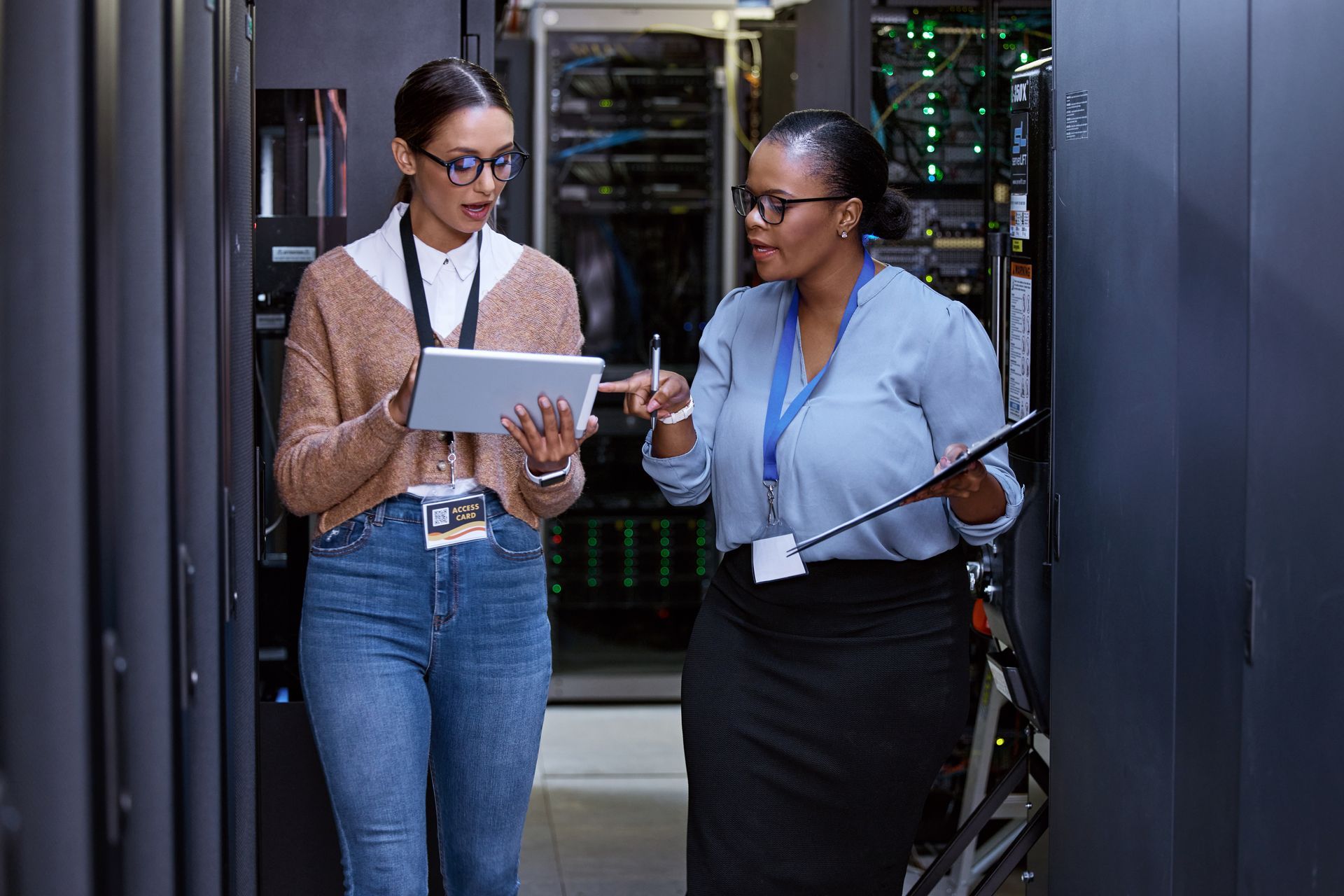 Two women in a server room, one holding a tablet, the other a clipboard, discussing data.