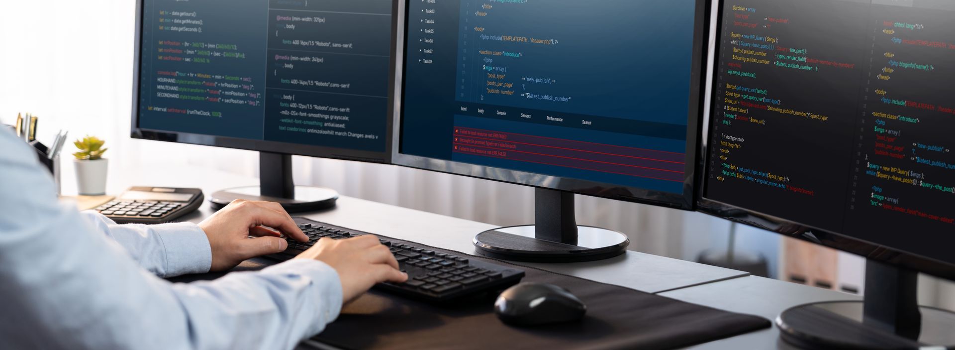 Person coding on a computer with three monitors displaying code. Keyboard and mouse on a desk.