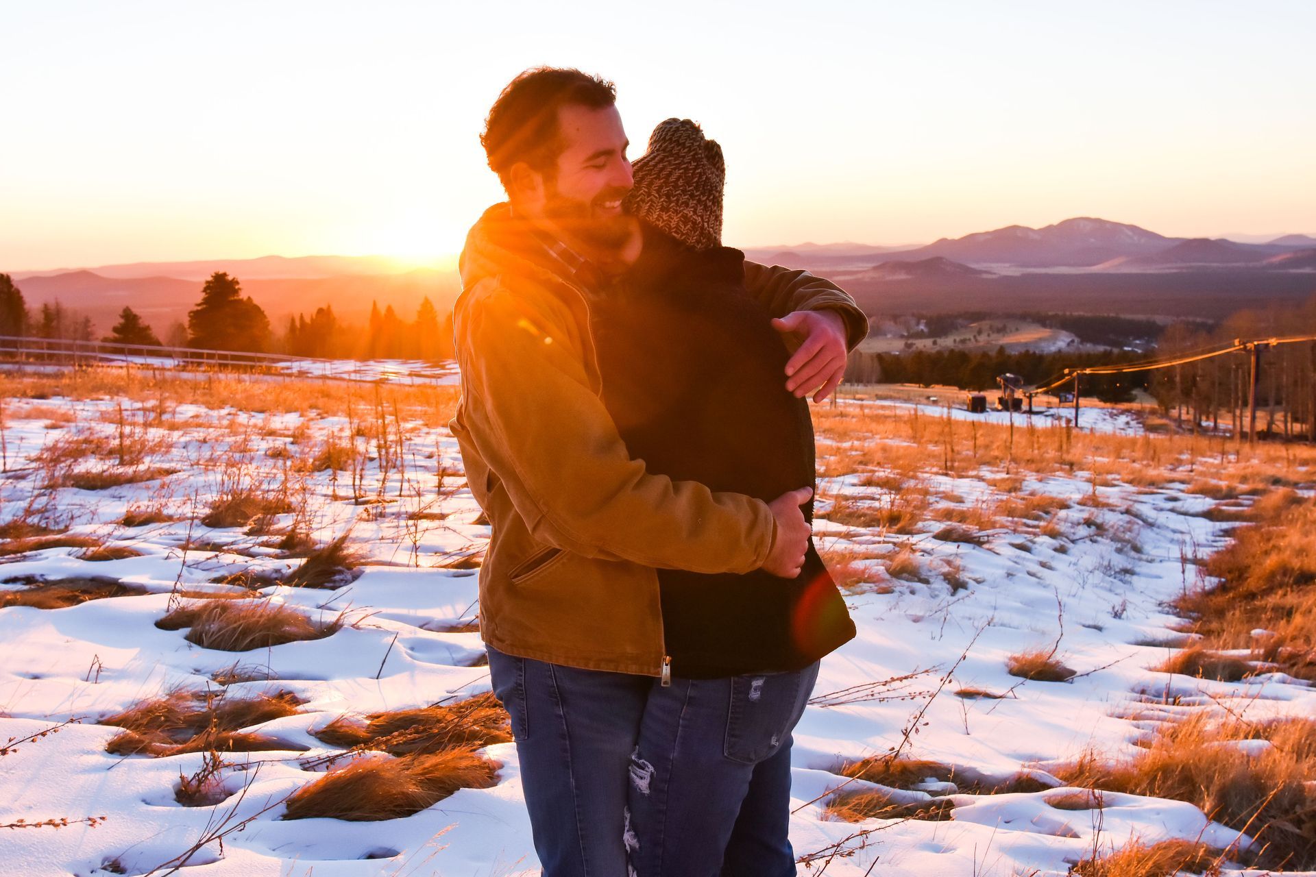 A man and woman are hugging in a snowy field at sunset.