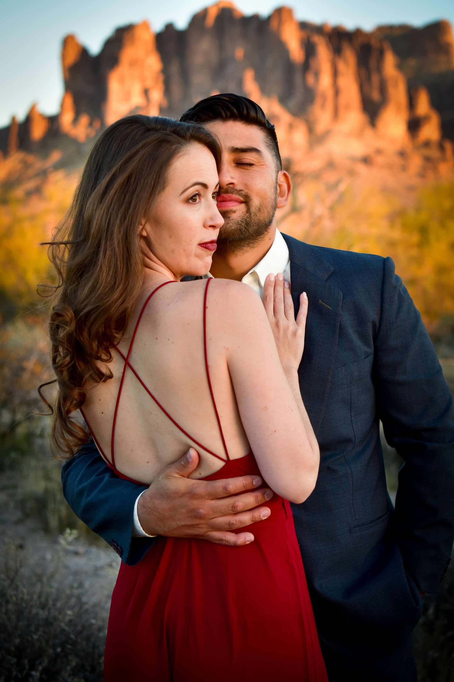 A man in a suit is hugging a woman in a red dress in front of a mountain.