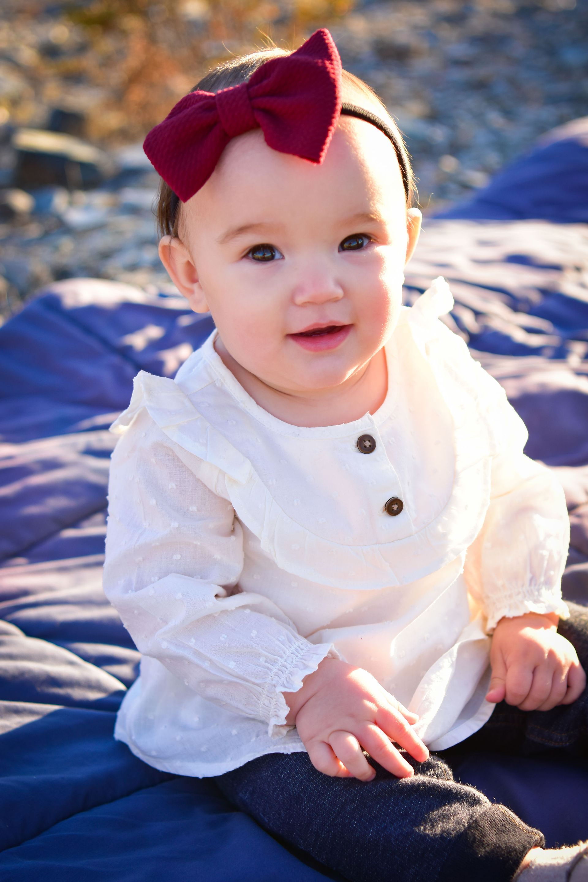 A baby girl wearing a white shirt and a red bow is sitting on a blue blanket.