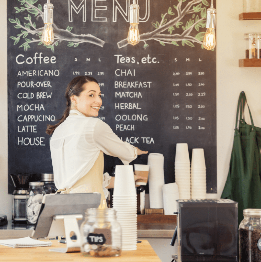 A barista points to a chalkboard menu in a coffee shop. Smiling, she's wearing an apron.
