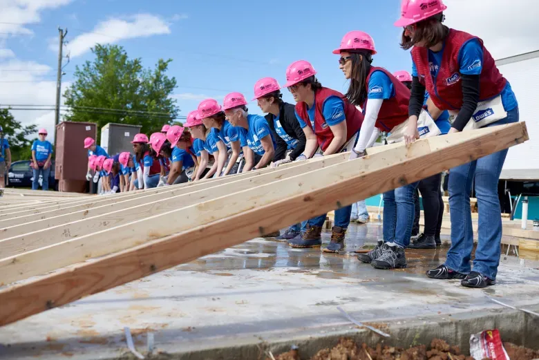 Women in pink hard hats and blue shirts lifting a wooden structure outdoors at a construction site.