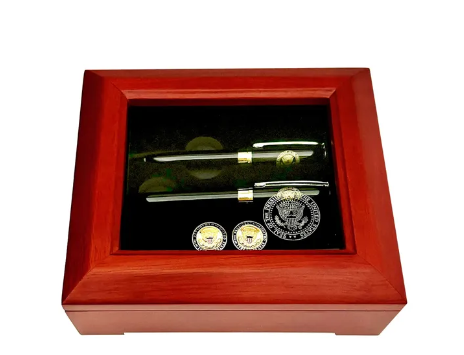 Display case with two pens and coins, all with gold accents, against a black backdrop, set in a red-brown wooden box.