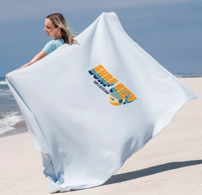 Woman on beach holding a light blue towel with a surf design.