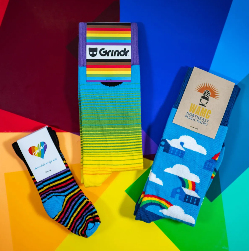 Three pairs of colorful socks on a rainbow background: Grindr socks, heart pride socks, and cloud/rainbow socks.