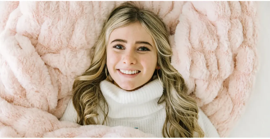 Blonde woman smiling, laying in a fluffy pink blanket, wearing a white turtleneck.