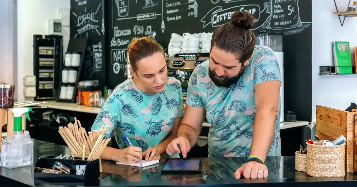 Two cafe workers in matching shirts, one writing, the other pointing at a tablet, behind a counter.