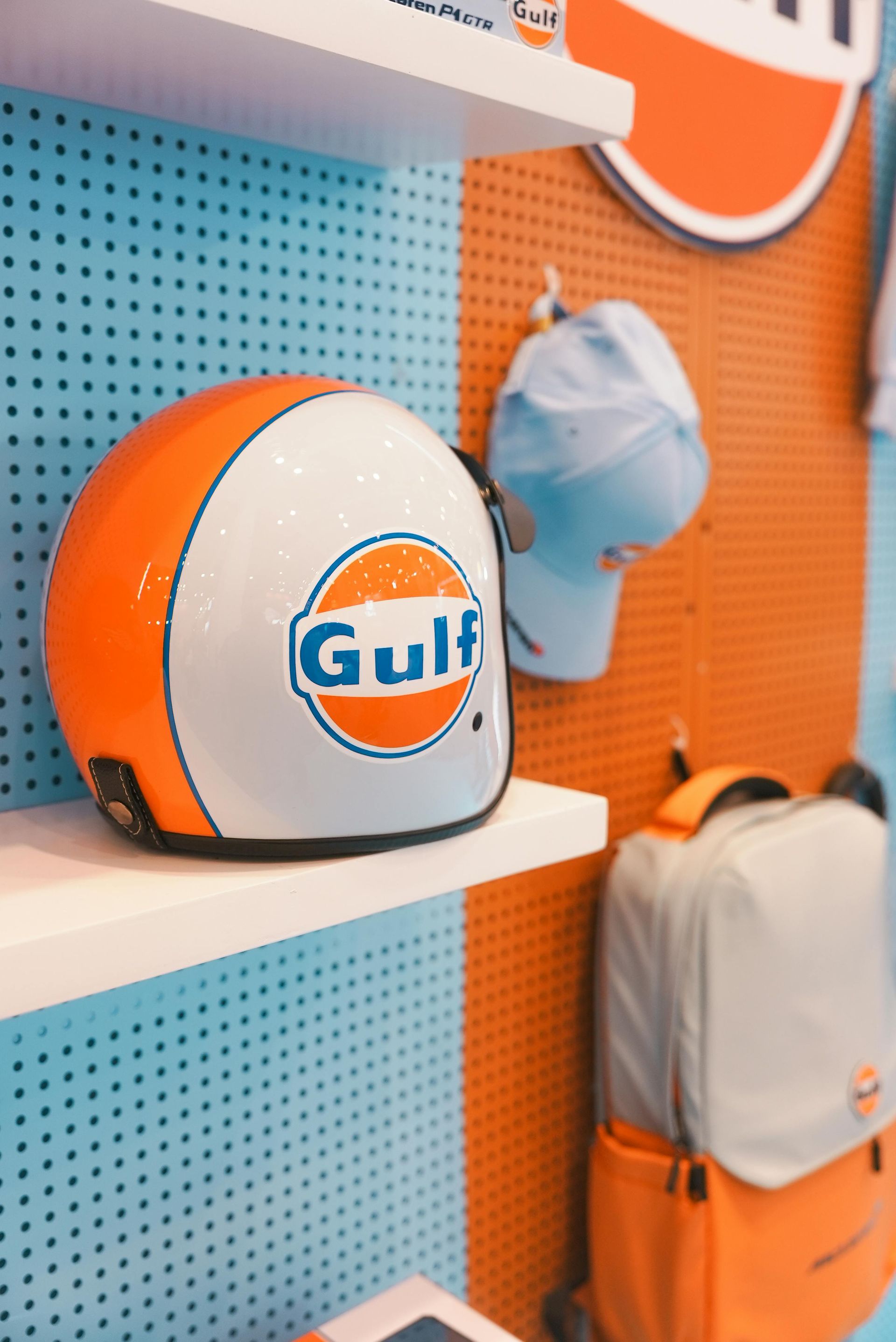 Gulf-branded motorcycle helmet on a shelf. Orange and white design. Blue and orange pegboard background.