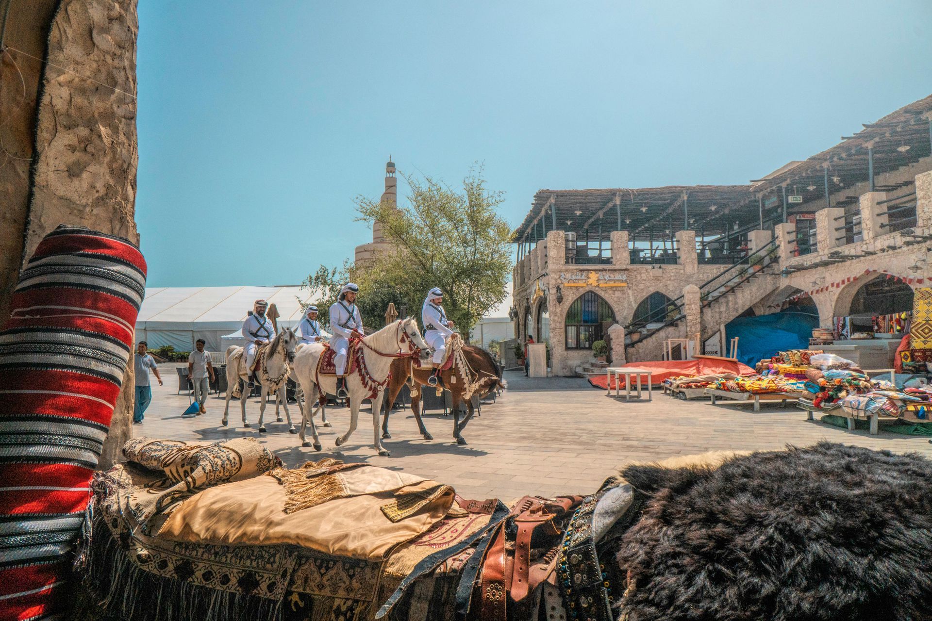 Men on camels in a courtyard with shops and a traditional building. Bright sky.
