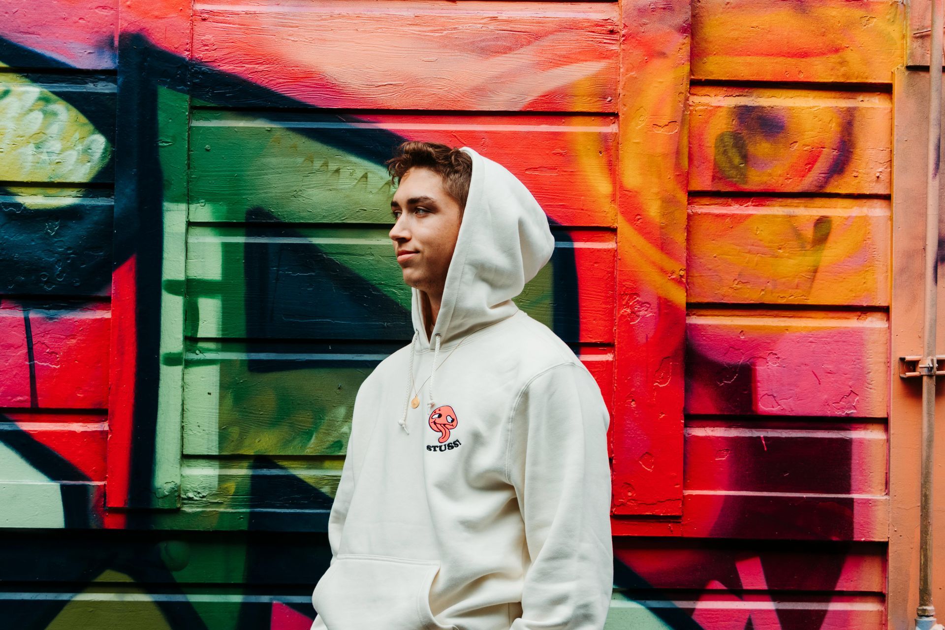 Man in a white hoodie stands in front of a colorful graffiti wall, looking to the side.
