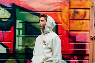 Man in a white hoodie stands in front of a colorful graffiti wall, looking to the side.