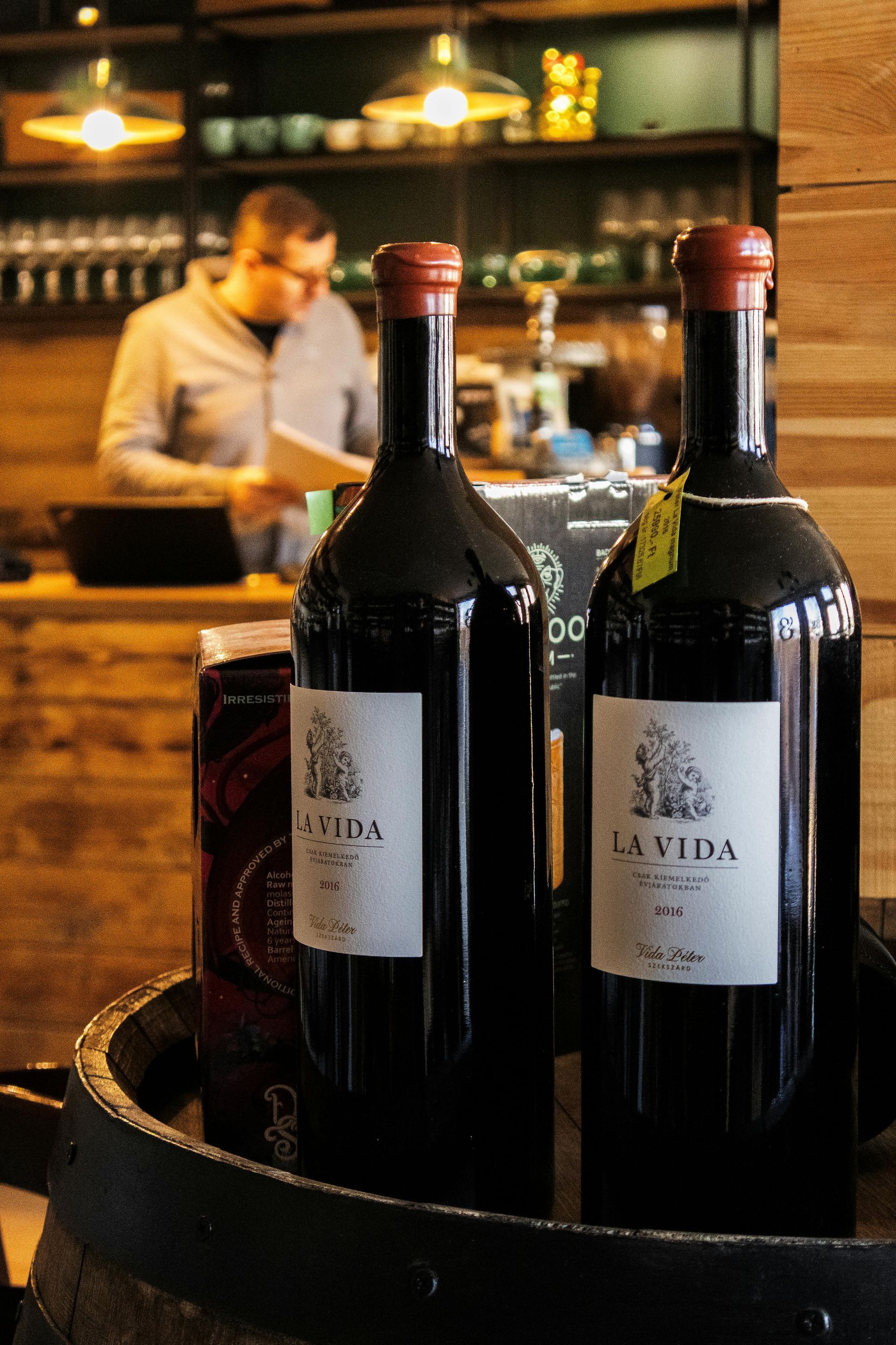 Two red wine bottles on a barrel, a person in the background reads.