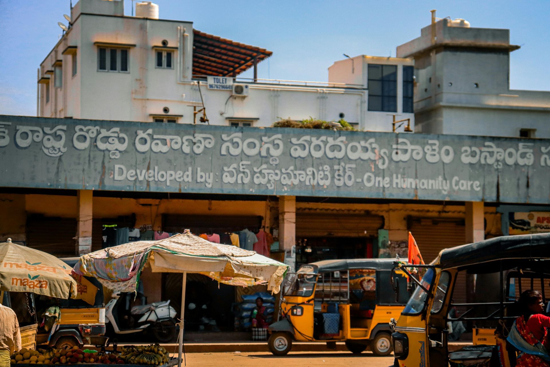 A bus station in India with street vendors and auto-rickshaws. The sign reads 
