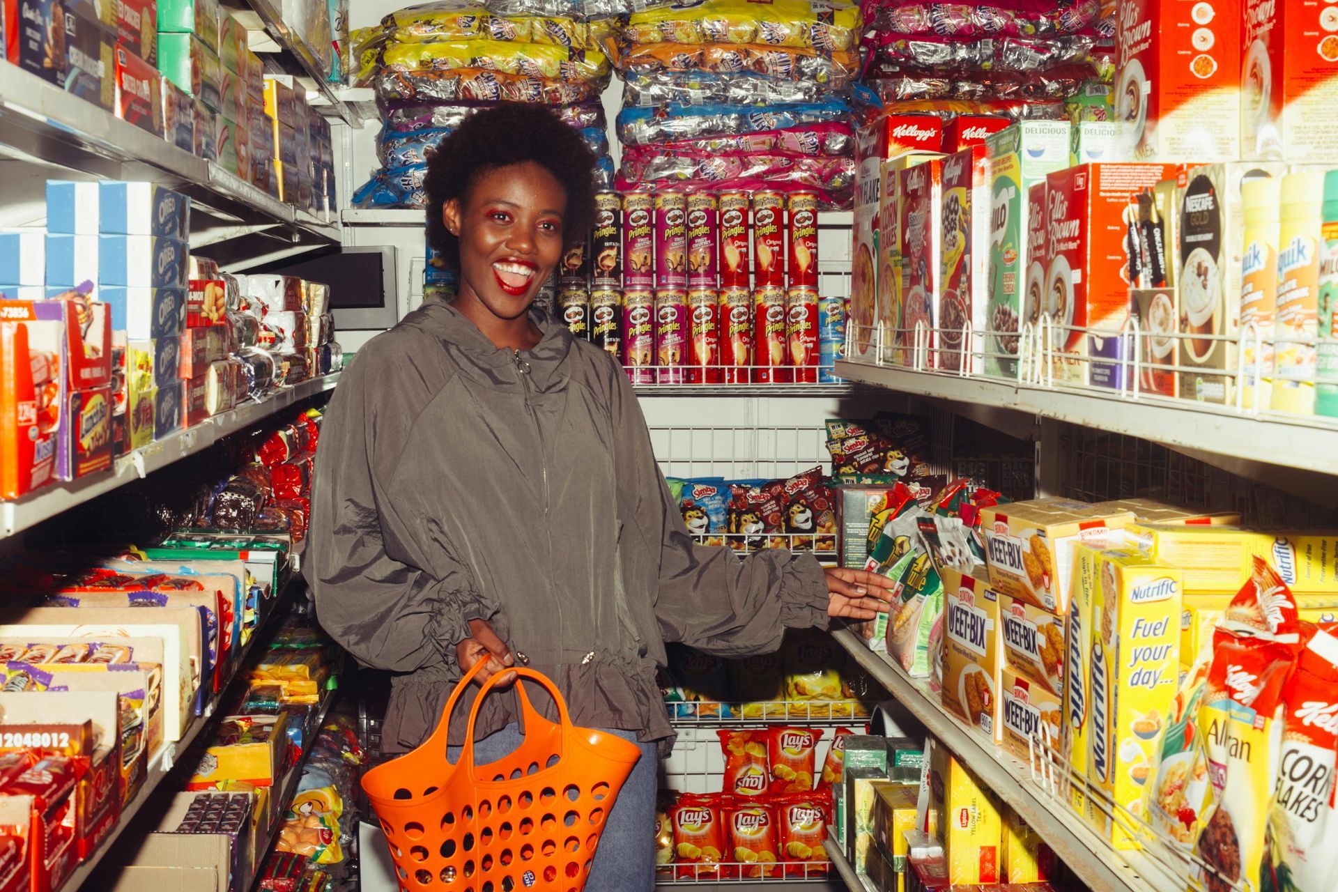 Woman in a grocery store aisle smiling, holding orange basket, surrounded by colorful food products.