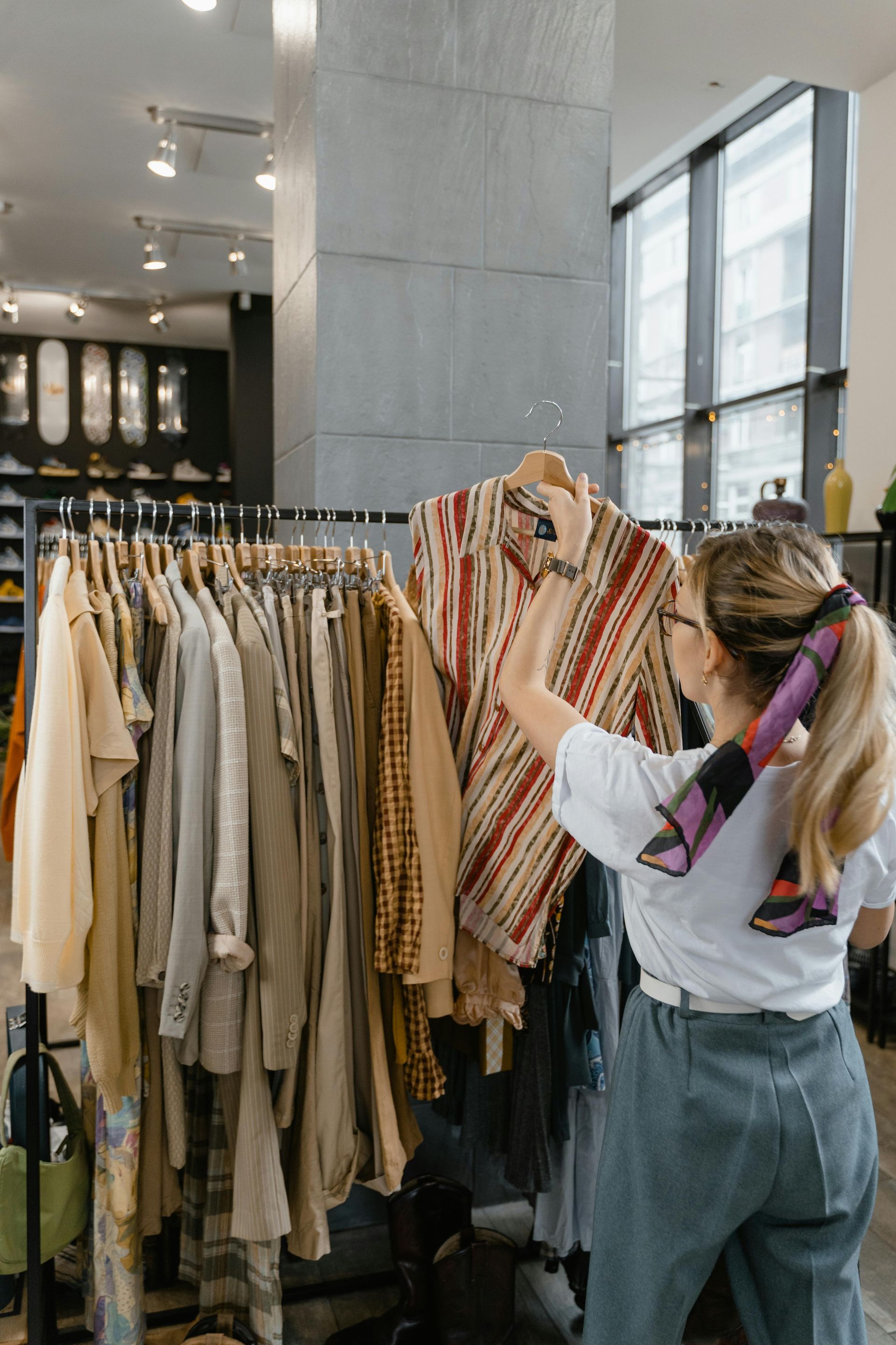 Woman shopping, looking at a striped shirt on a rack of clothing inside a store. She wears glasses and a scarf.