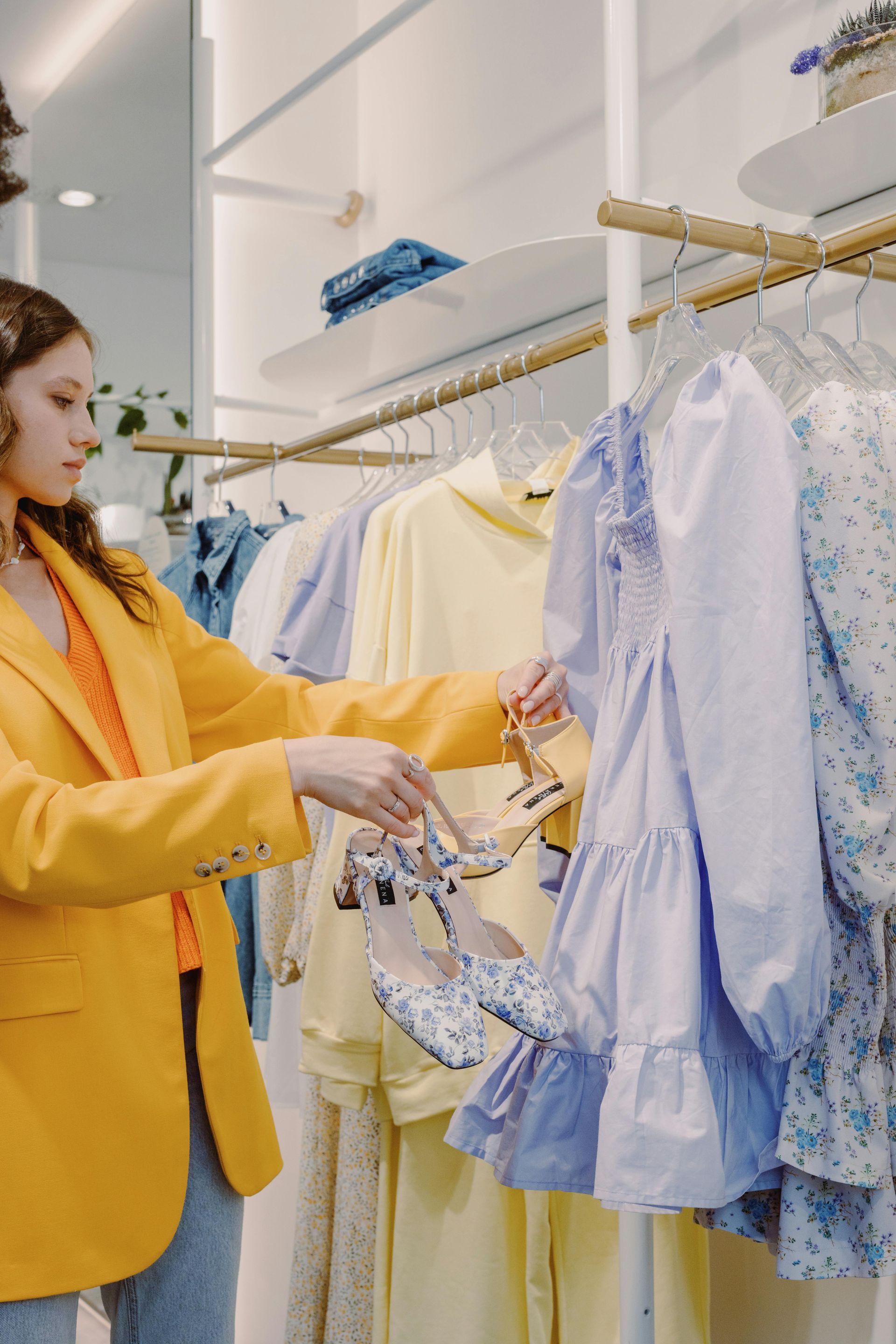 Woman in yellow blazer and jeans holding shoes, looking at a lavender dress in a boutique.