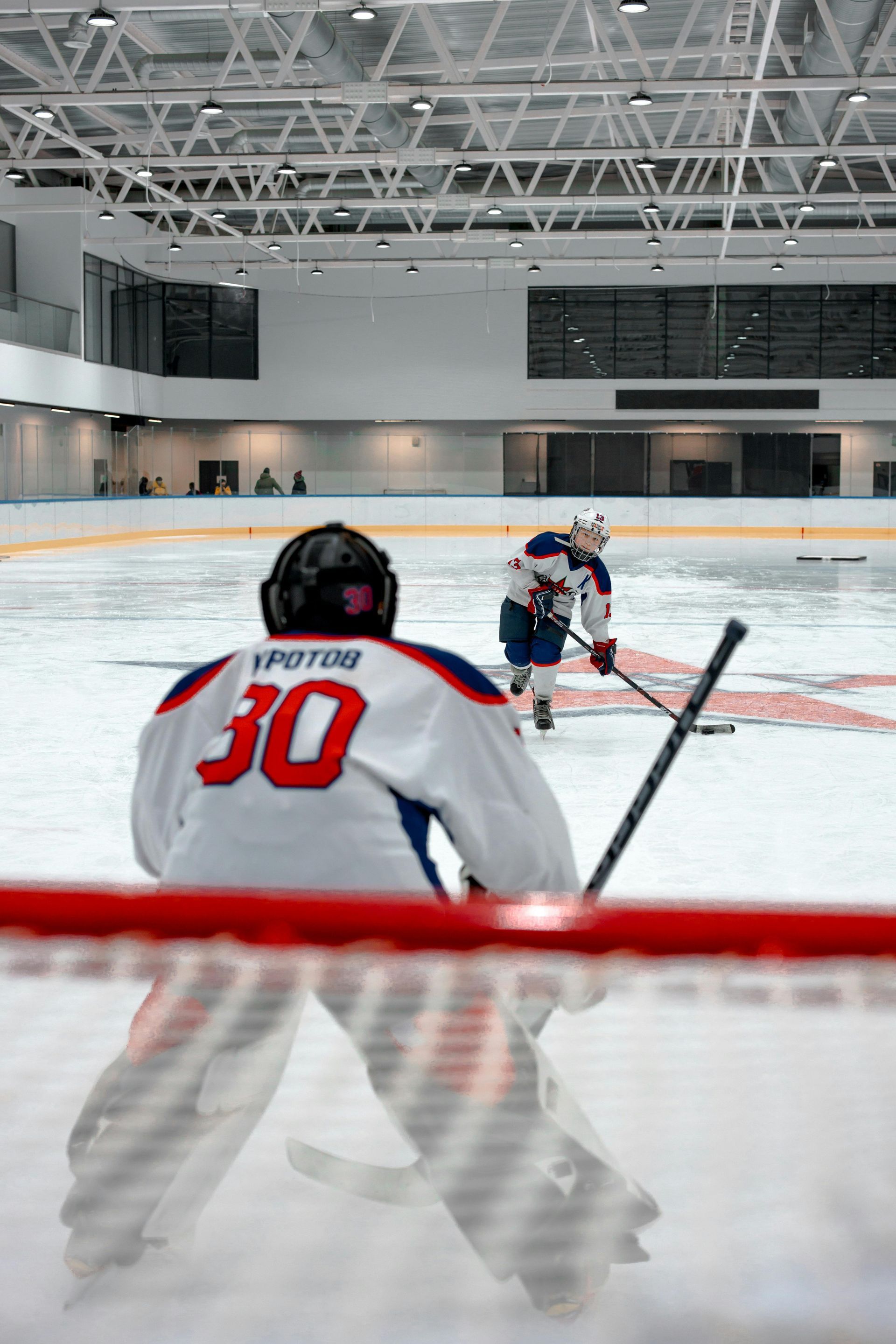 Hockey goalie in net facing a player on ice rink. Number 30 on goalie's back, stick in hand, aiming.