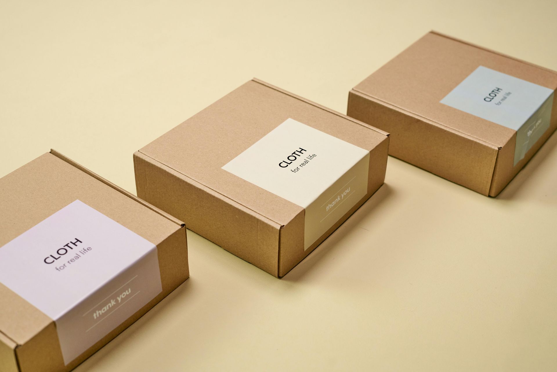 Three square, brown cardboard boxes with pastel labels arranged on a light-yellow surface.