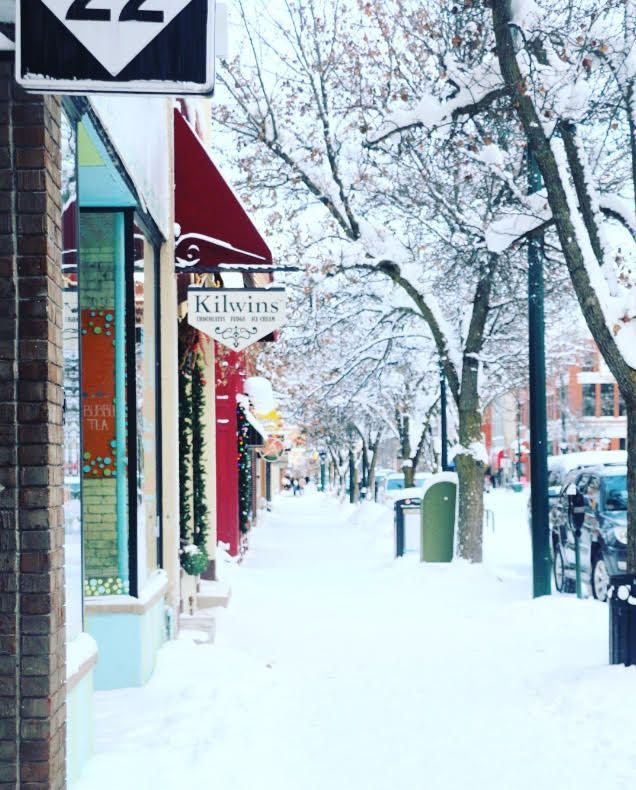 Snowy street scene, buildings with colorful storefronts, sign for Kilwins, trees covered in snow.