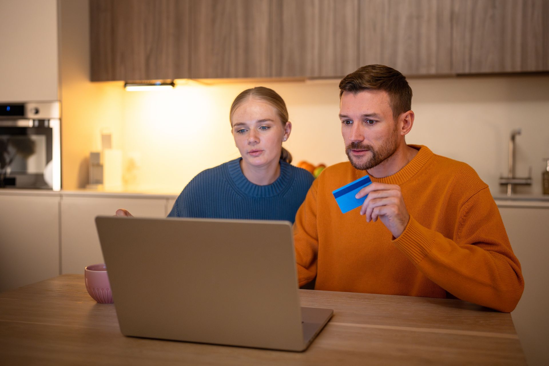 A couple looking at a laptop, man holding a blue credit card, in a kitchen setting.