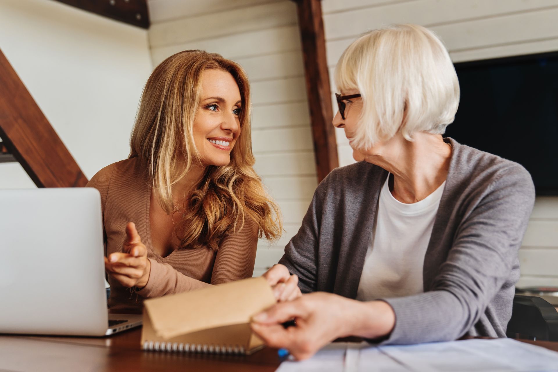 Woman showing document to older person next to laptop. Both smiling in a wood-paneled room.
