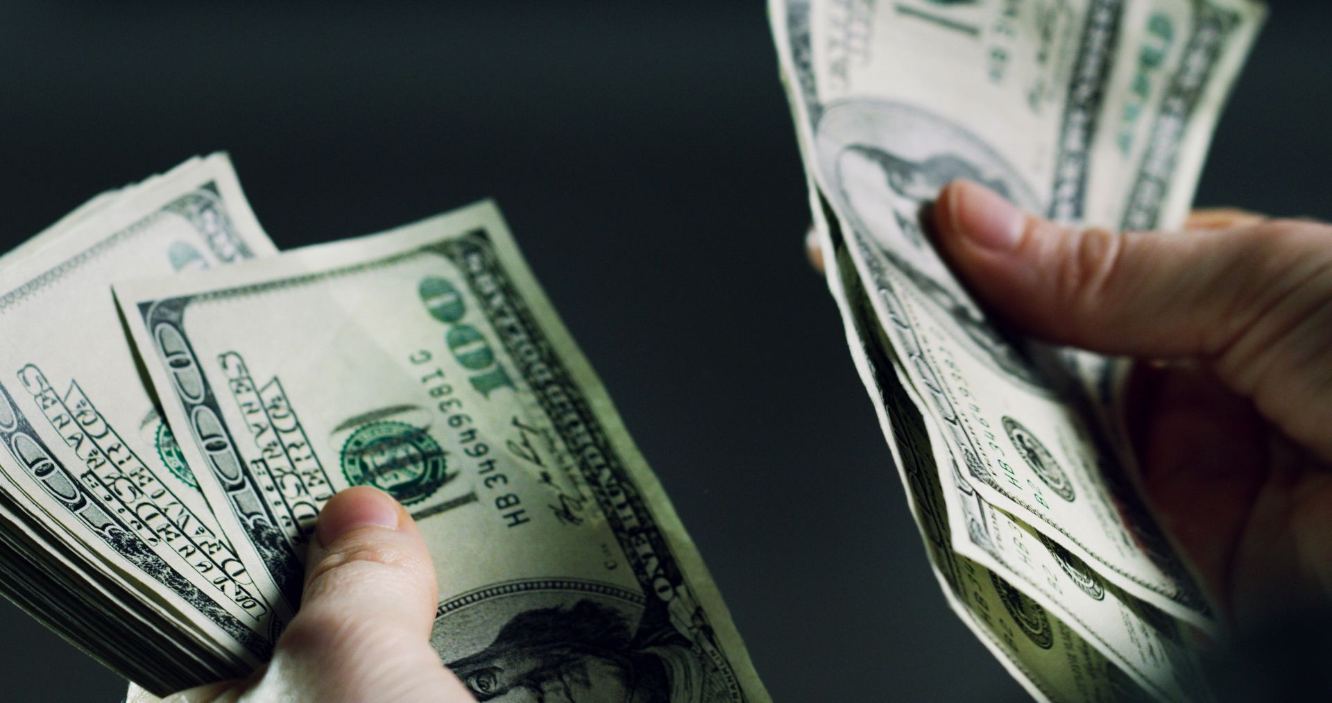 Hands holding and counting stacks of one-hundred dollar bills against a dark background.