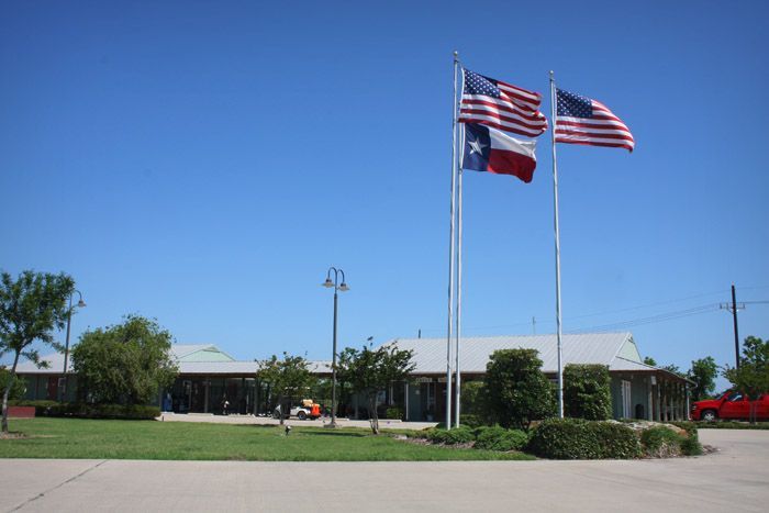 Building with three flagpoles: American flag, Texas flag, and another American flag flying against a blue sky.