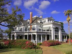 White mansion with pillars, balcony, and awnings on a sunny day with blue sky and palm tree.