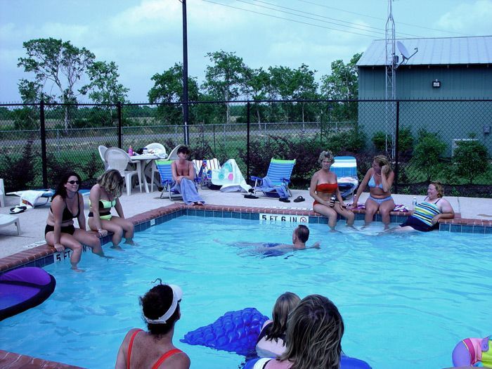 People in swimwear sit around a pool, with others swimming. Cloudy sky, fence, and building in the background.